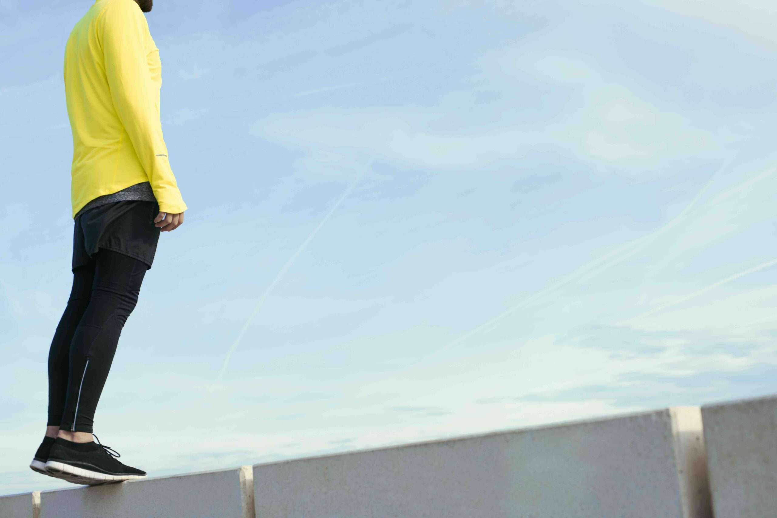 Sportif en coupe-vent jaune prenant une pause, les mains croisées, debout sur fond de ciel. Homme en forme se reposant après une séance d'entraînement en plein air, coureur se reposant après un entraînement de fitness.