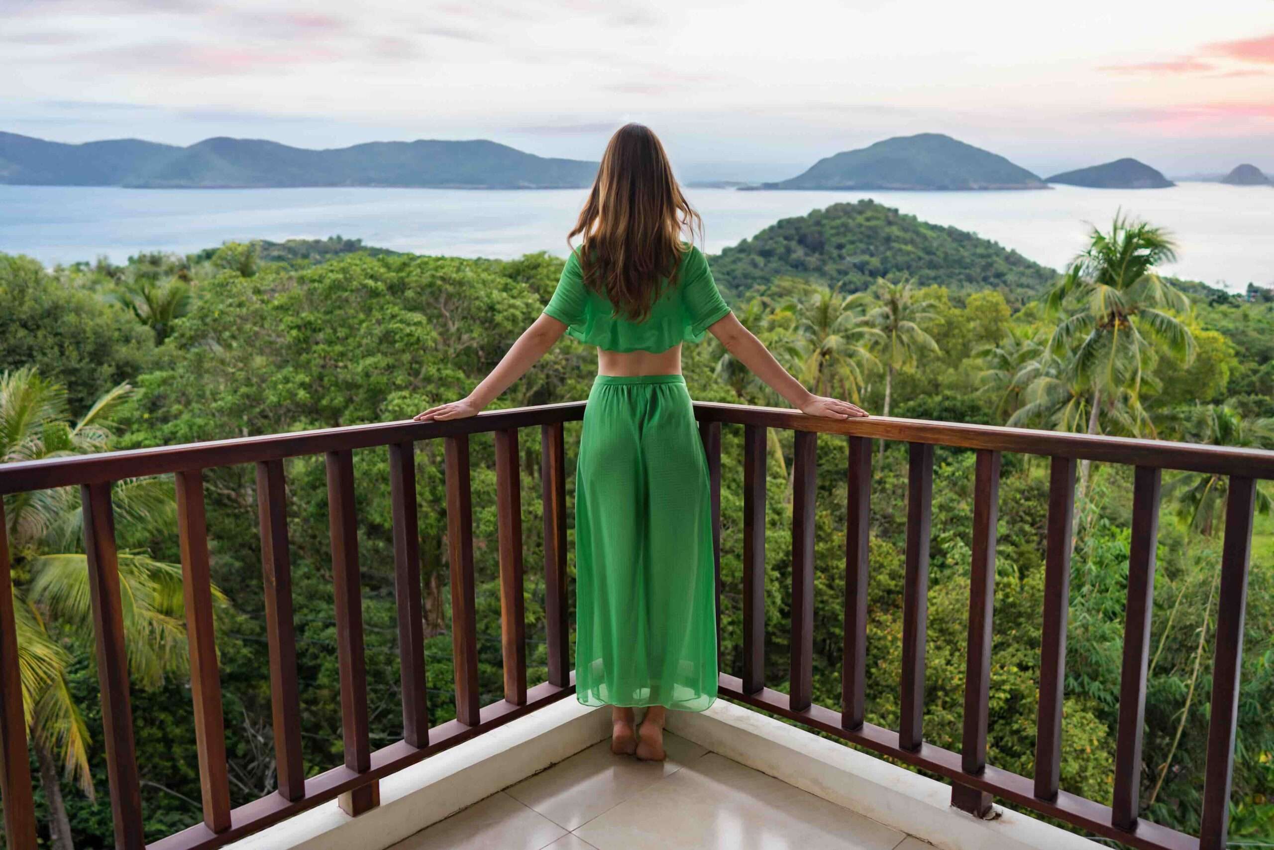Une jeune femme en robe verte se tient sur un balcon dans un pays tropical et regarde vers la jungle